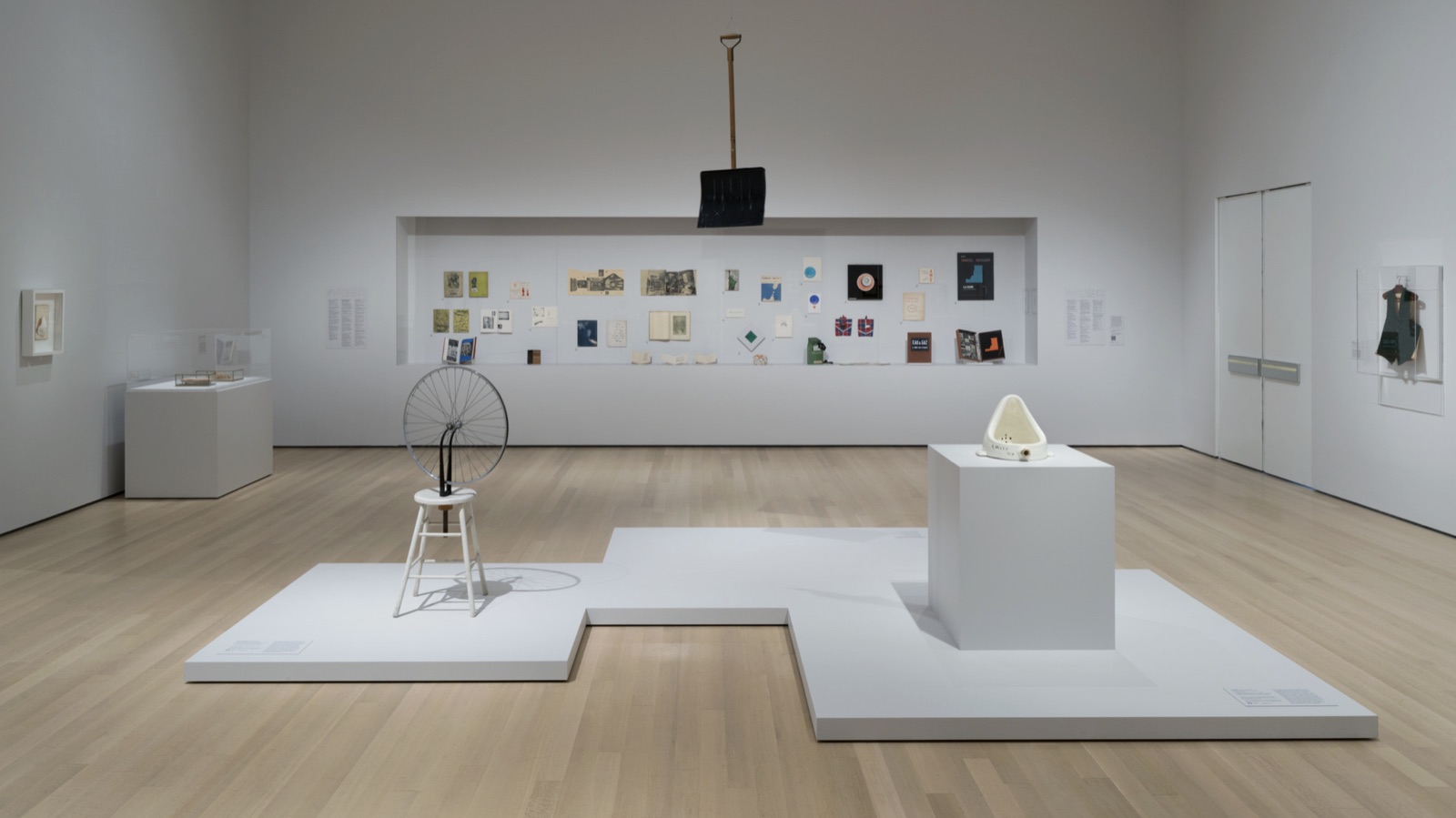Installation view of a white gallery: Bicycle Wheel mounted on a stool, Fountain on a pedestal, a snow shovel suspended from the ceiling, a wall of small printed works behind