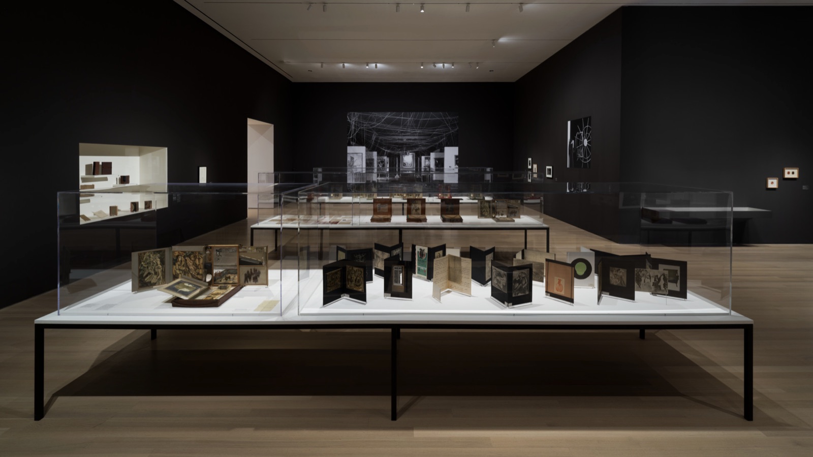 A dark gallery with vitrines holding a dozen open Box in a Valise editions, a large photographic mural on the far wall