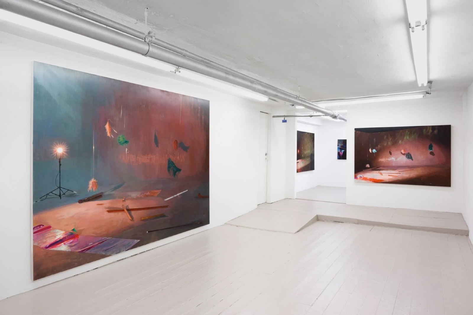 Theta's basement gallery: white walls, a galvanized pipe and fluorescent tubes along the ceiling, a large painting of a lit stage with a lamp and hanging feathers at left, two more paintings beyond